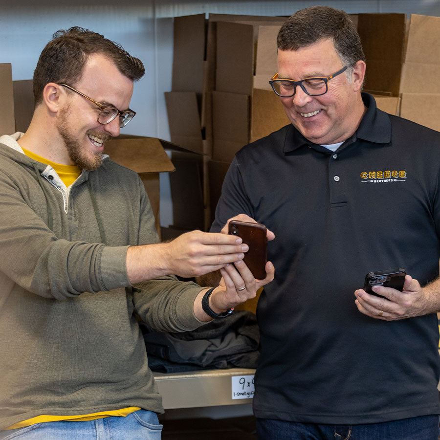 Cheese Brothers Eric and Gene looking at a phone together in a warehouse setting with boxes in the background