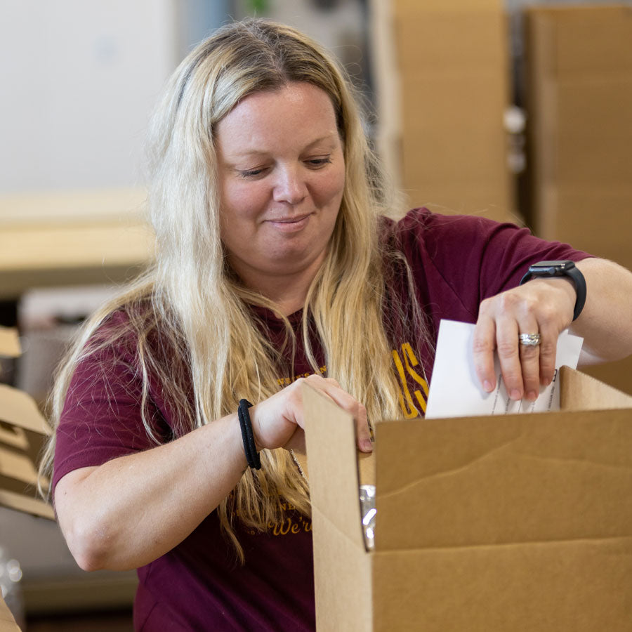 Woman in maroon t-shirt with yellow Cheese Brothers logo packing a cardboard box in a warehouse.