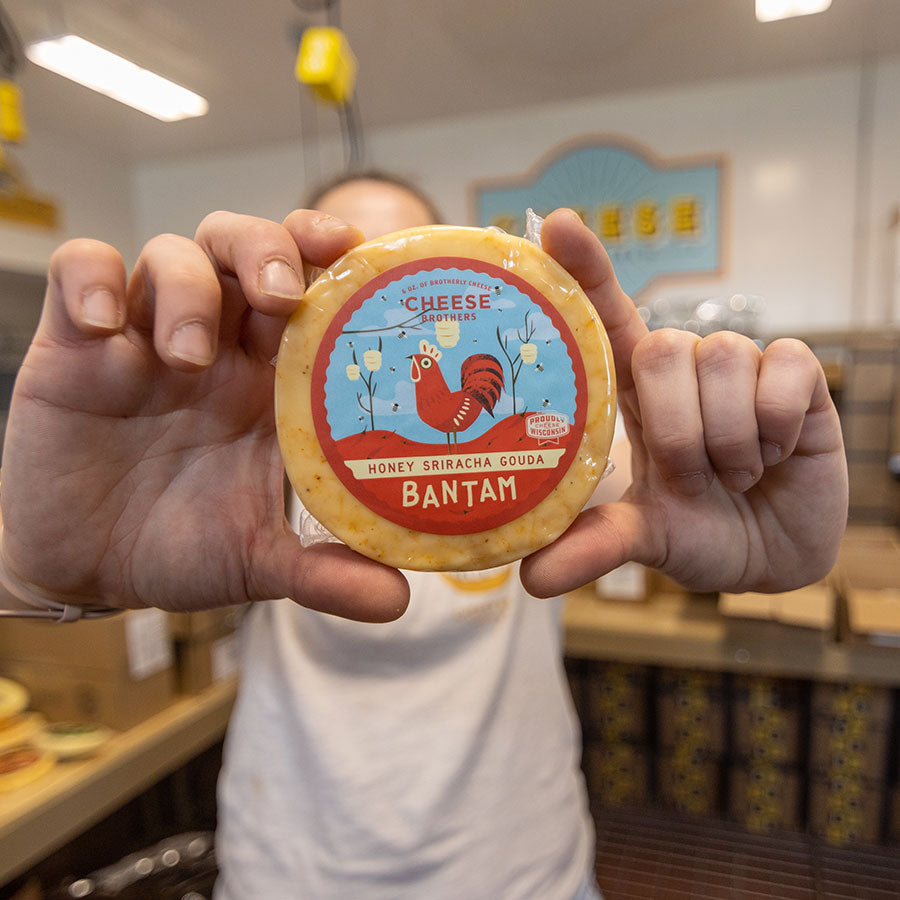 Person holding a pack of Cheese Brothers Bantam Honey Sriracha Gouda Cheese in a store setting