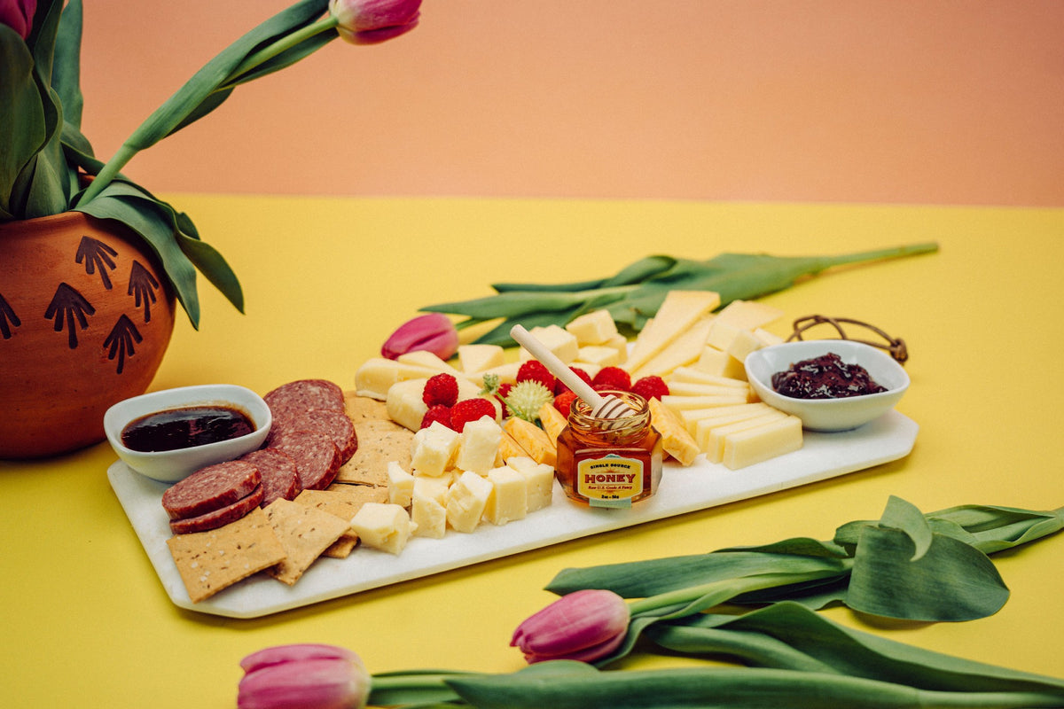 A plate made from the Mother&#39;s Day charcuterie box with a variety of artisan cheeses, honey, and jams next to flowers.