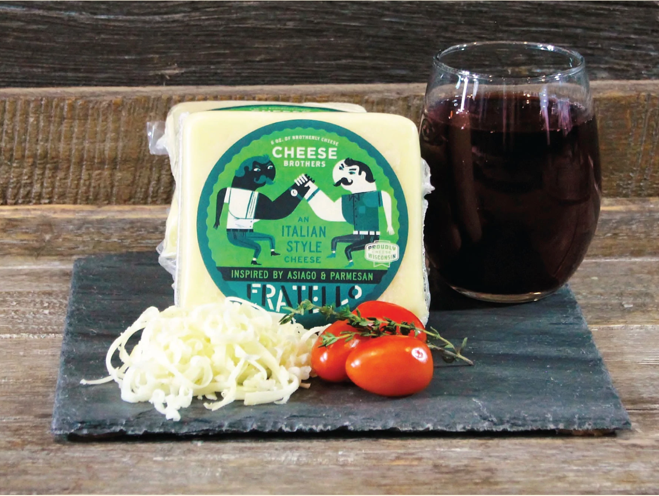 Package of Fratello asiago and parmesan cheese blend on a black slate next to tomatoes and a glass of red wine. 