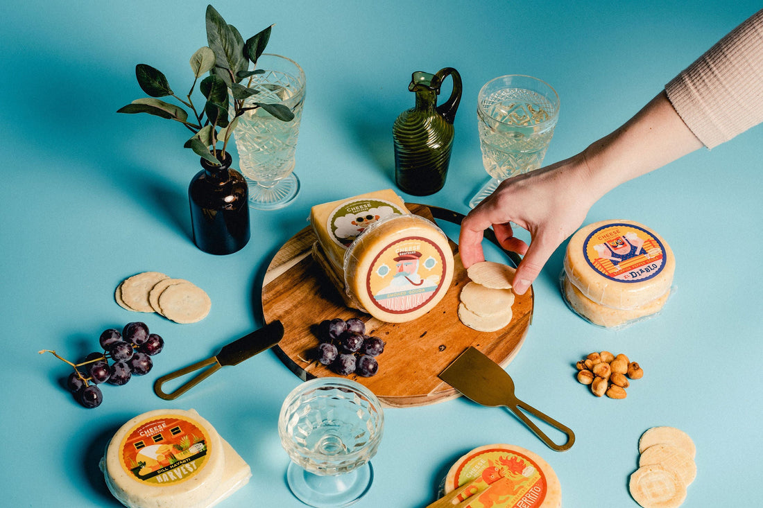 Hand picking up cracker from charcuterie board with grapes and variety of artisan Wisconsin cheese packages. 