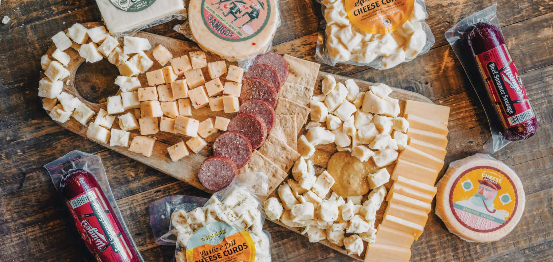 Charcuterie board with a variety of artisan Wisconsin cheeses, curds, sausages and mustard surrounded by packages. 