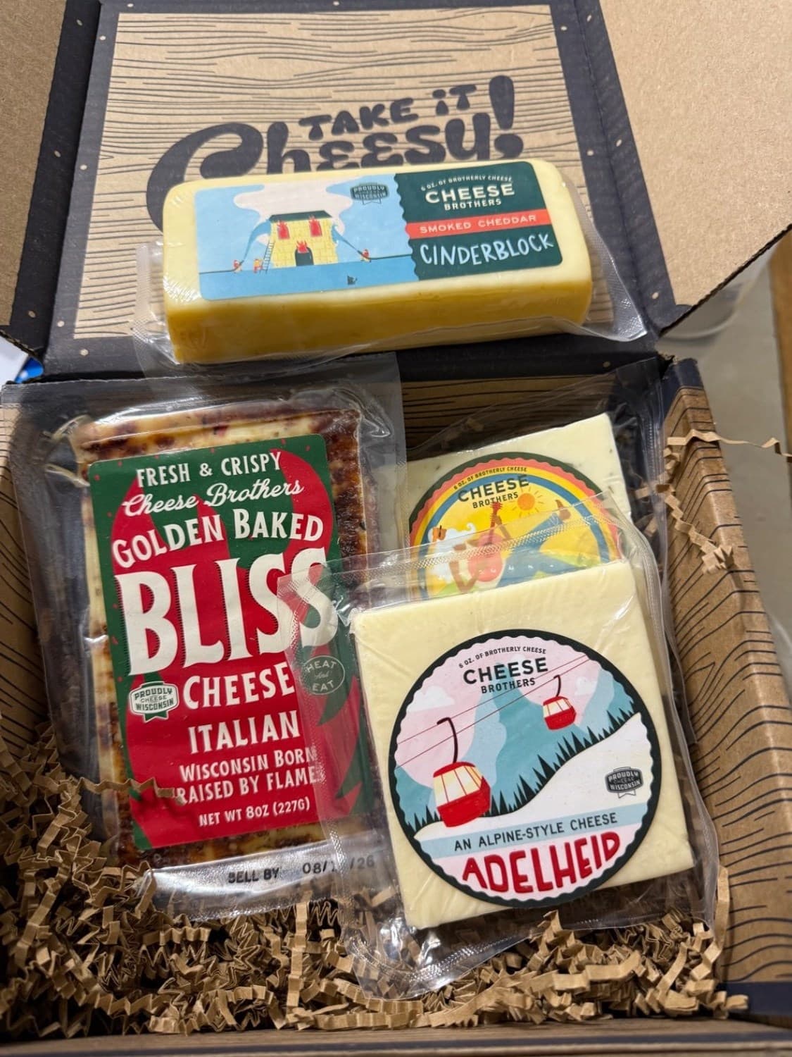 Assorted cheese packages including 'Bliss' and 'Adelheid' in a box with 'Take it Cheesy!' text.