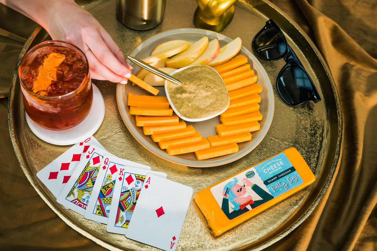 Block of Pretty Boy sharp Wisconsin cheddar next to playing cards, a cocktail, and charcuterie plate. 