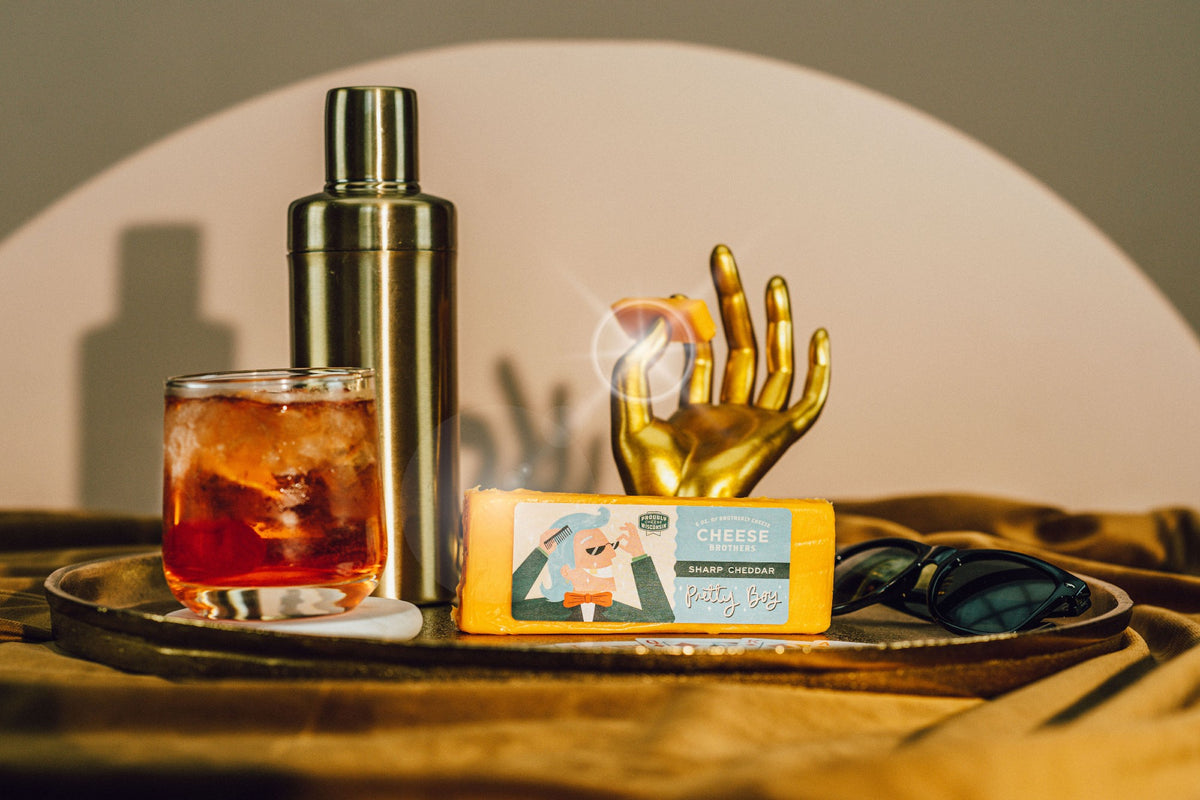 Block of Pretty Boy sharp Wisconsin cheddar next to cocktail and golden hand holding a slice of cheese.