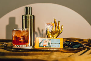Block of Pretty Boy sharp Wisconsin cheddar next to cocktail and golden hand holding a slice of cheese.