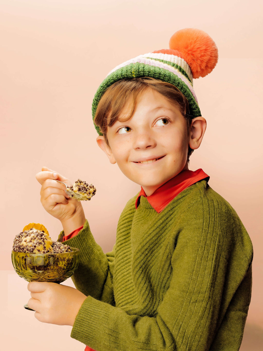 Child wearing a green sweater and colorful winter hat holding a glass bowl full of chocstasy dessert cheese.