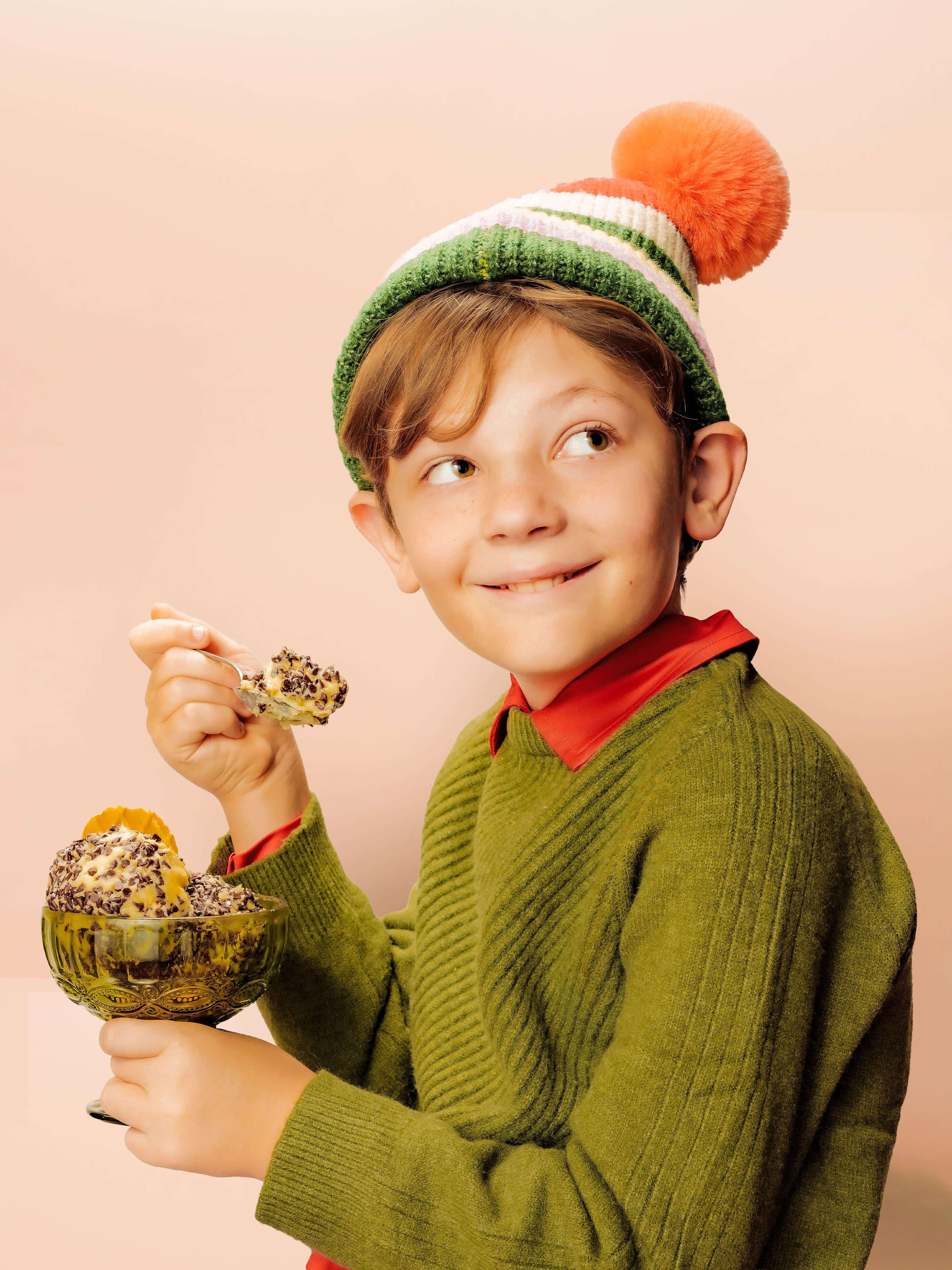 Child wearing a green sweater and colorful winter hat holding a glass bowl full of chocstasy dessert cheese.