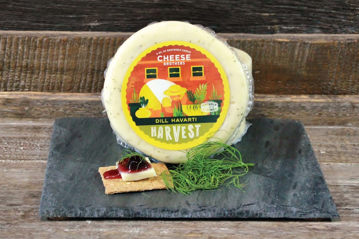 Package of dill Havarti Harvest artisan cheese on a black slate behind cheese and crackers and dill sprig.