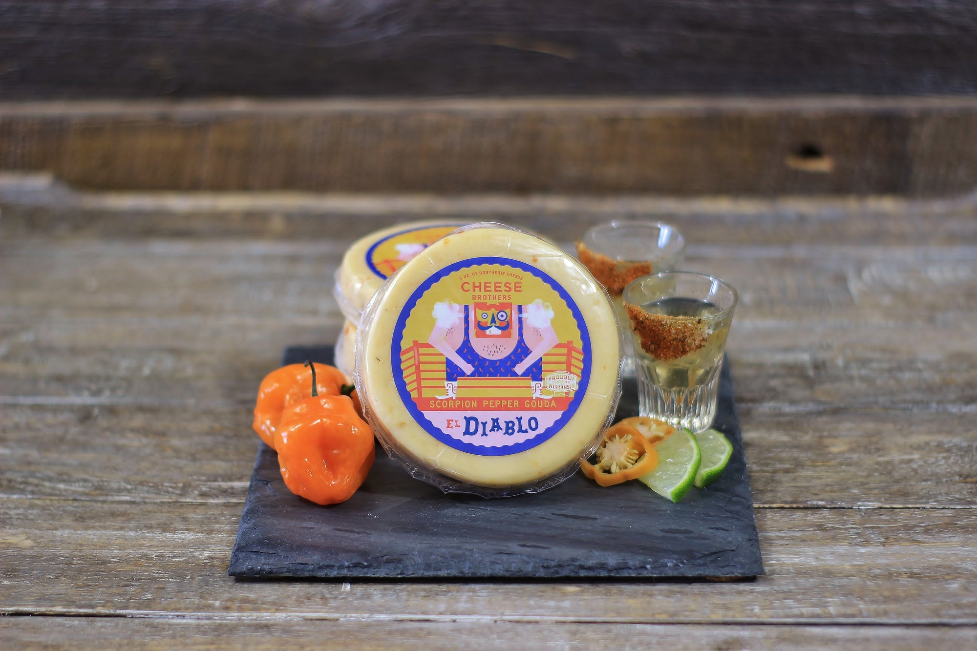 Package of El Diablo scorpion pepper gouda cheese on a black slate with peppers and shots of tequila. 