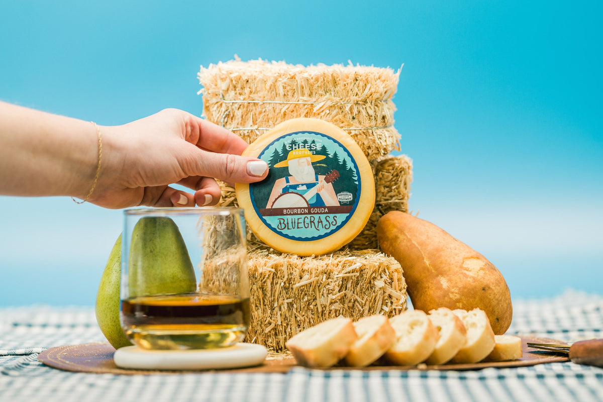 A package of Bluegrass bourbon gouda on stacks of hay next to glass of bourbon and bread. 