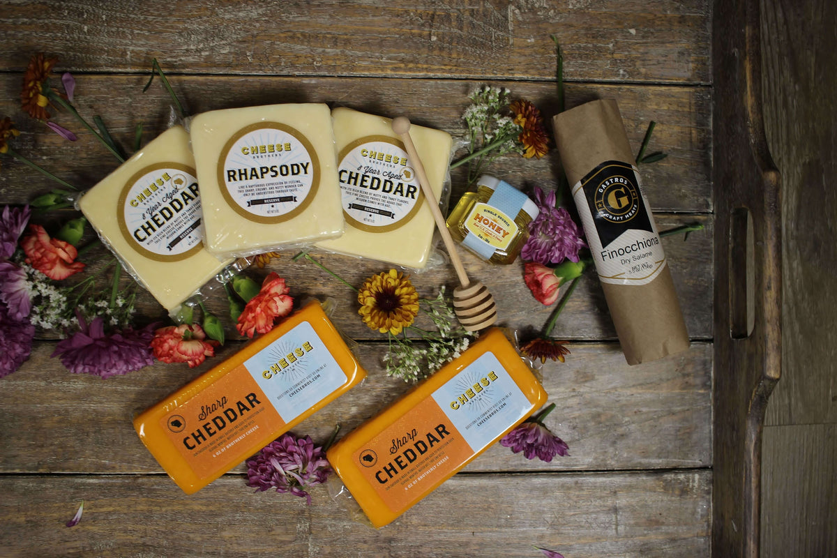 Five packages of gourmet Wisconsin cheese, honey, sausage, and flowers on wooden tray. 