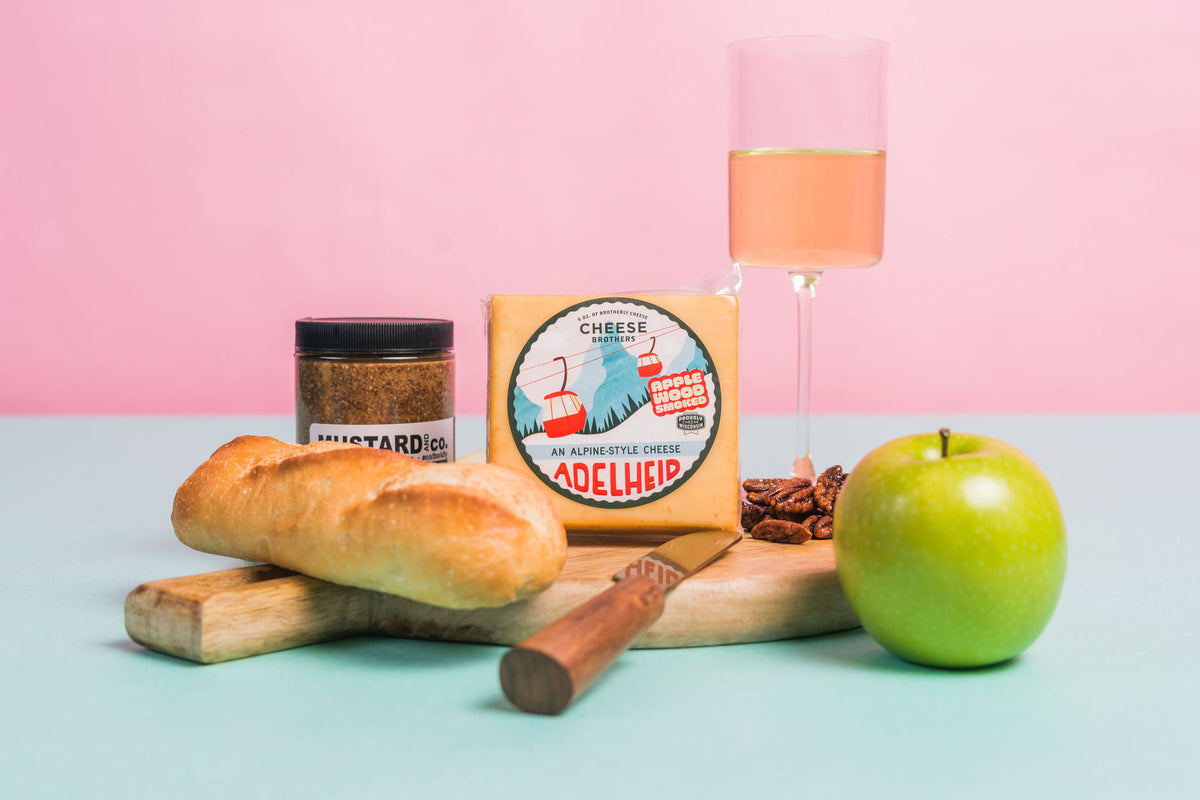 Package of Adelheid cheese on a charcuterie board with nuts, mustard, bread, a green apple, and a glass of wine. 
