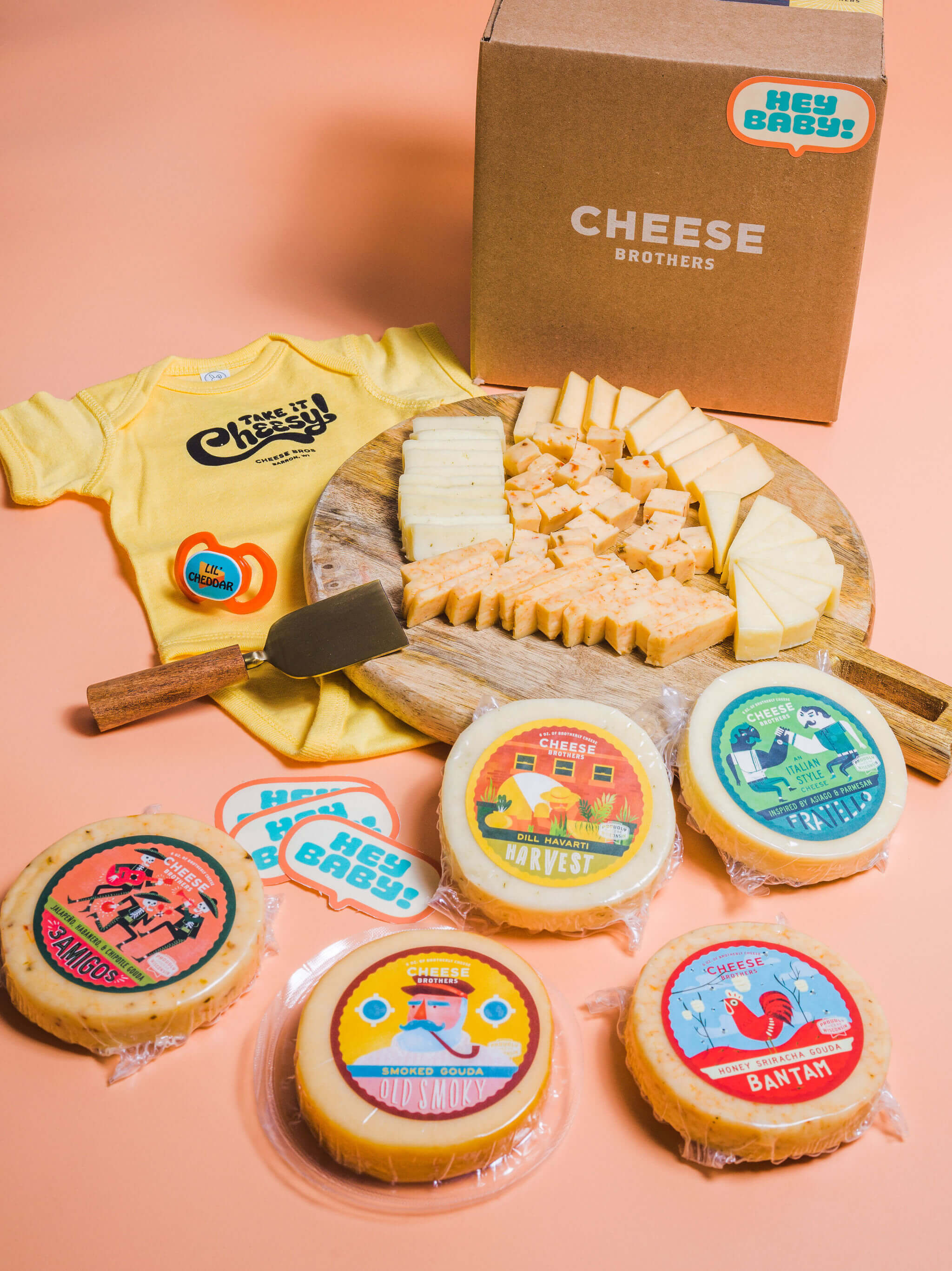 A variety of artisan cheese packages, a charcuterie board, Cheese Brothers baby onesie, and delivery box.  