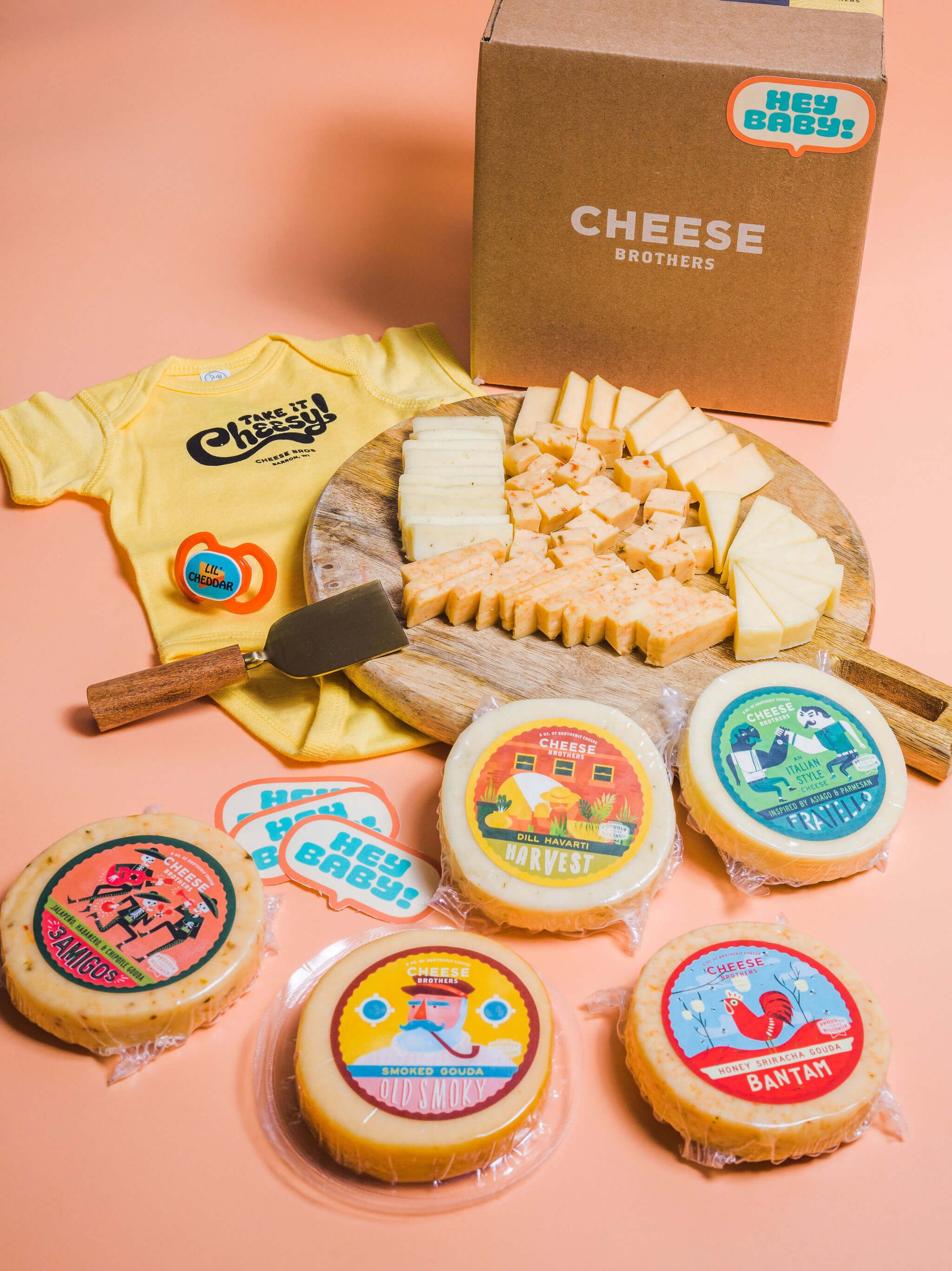 Seven varieties of artisan Wisconsin cheese with a Cheese Brothers baby onesie in front of a box. 