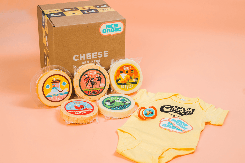 Seven varieties of artisan Wisconsin cheese with a Cheese Brothers baby onesie in front of a box. 