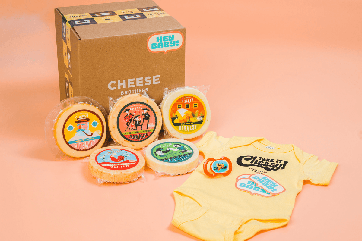 Seven varieties of artisan Wisconsin cheese with a Cheese Brothers baby onesie in front of a box. 