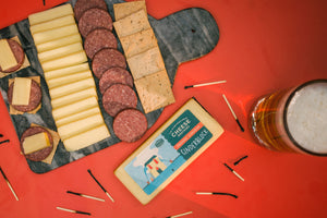 Block of cinderbloked smoked cheddar cheese next to burn matchs and a meat and cheese black charcuterie board. 