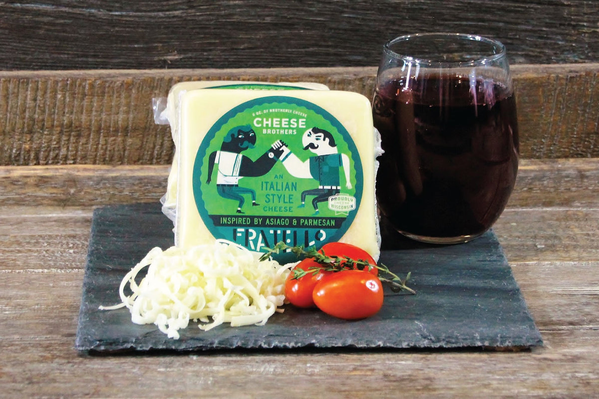 Package of Fratello asiago and parmesan cheese blend on a black slate next to tomatoes and a glass of red wine. 