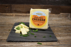 Package of artisan green onion Wisconsin cheddar on a black slate with chopped green onions and cheese slices. 