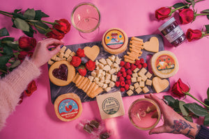 Hands picking from a black charcuterie board with packages of gourmet cheeses and caramel bites. 
